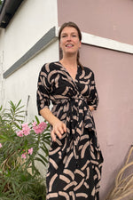 Woman in the long wrap Letsa Dress in the black & tan batik print, Cantaloop standing outside, looking elegant & stylish.