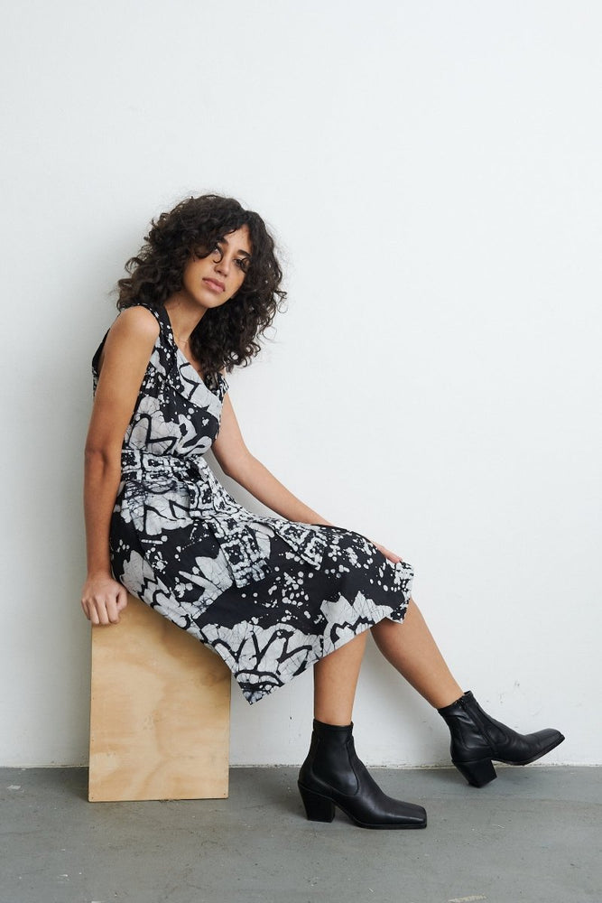 Duplo Dress in 2 Party System print, model seated on wooden bench, black ankle boots, against white wall.