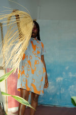 Short Helia Dress with orange-blue abstract print, in front of blue wall with leafy plant.