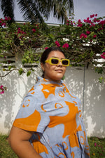 Outdoor shot of person wearing sunglasses in the Helia Dress with orange and blue print,  next to pink flowers