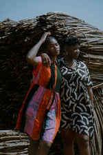 Two models in dresses by a rustic stick shelter, one in colorful Carmine print, the other in black and tan Cantaloop print.
