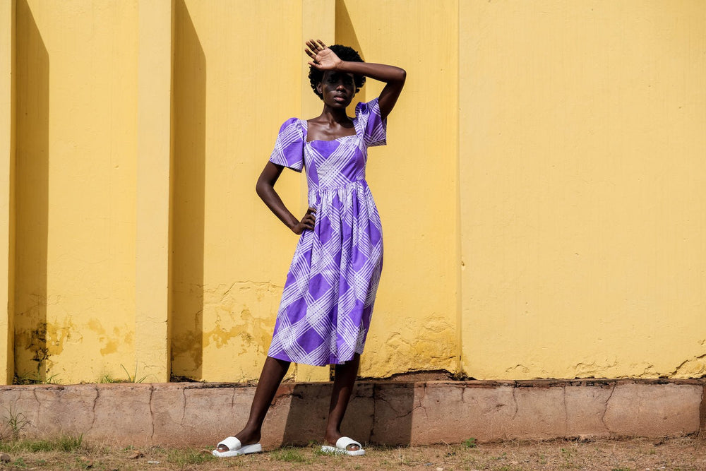 A woman in the Abeo Dress in Eostre, a purple and white hand-batiked print, posing in front of a vibrant yellow wall.