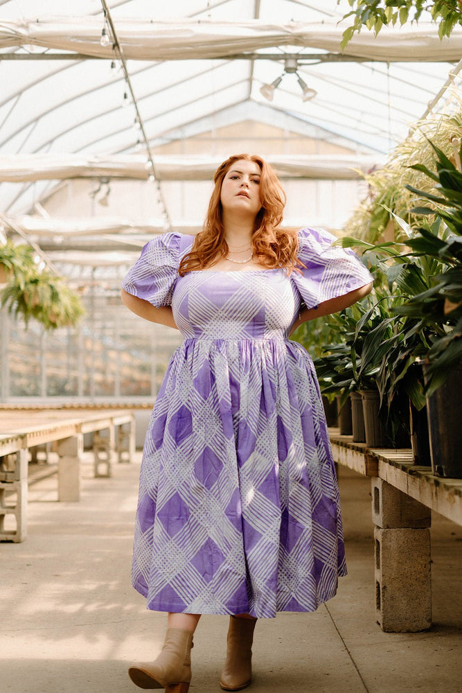 Woman in the Abeo Dress in Eostre, a purple and white print, standing in greenhouse surrounded by lush green plants.