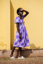 A woman in a hand-batiked purple and white Abeo dress posing in front of a vibrant yellow wall.