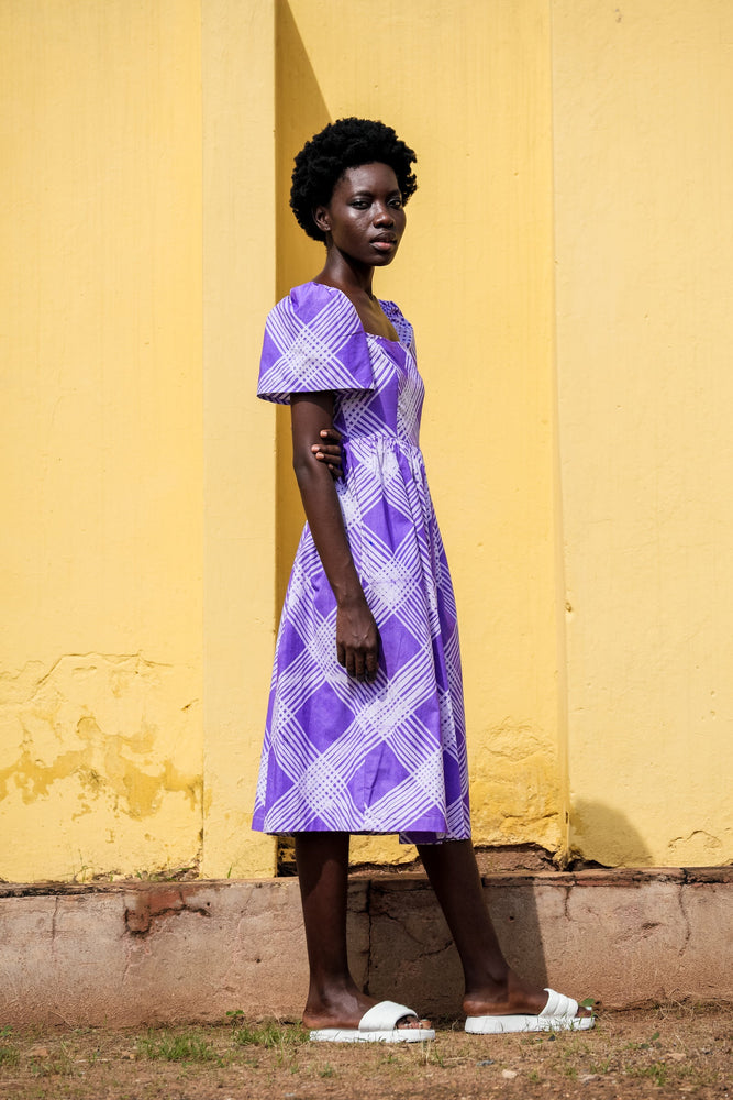Stylish lady in a purple dress stands confidently in front of a bright yellow wall. Hand dyed cotton with puffed sleeves.