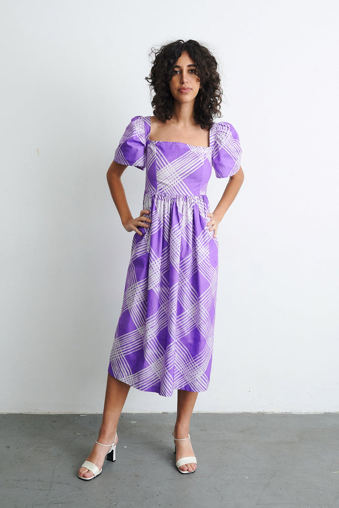 A woman wearing a hand dyed purple Abeo dress made of 100% cotton with puffed sleeves, rouching at back, full skirt, pockets.