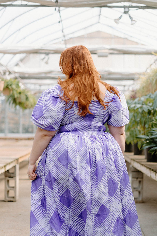 Back view of a woman in the Abeo Dress in Eostre, a purple and white print dress, strolling through lush greenhouse.