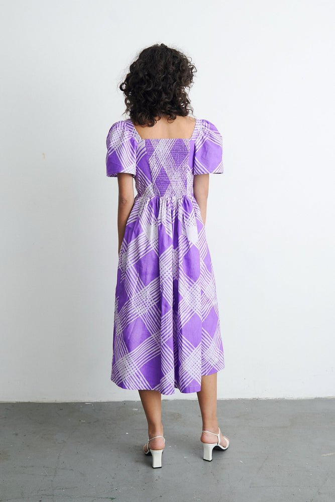 Back view of a trendy woman wearing the Abeo dress in the purple and white plaid print, Eostre. Hand-batiked cotton fabric.