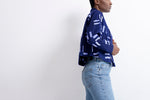 Abiba Jacket in blue with intricate white patterns, styled with light jeans, capturing a poised turn on a white stage.