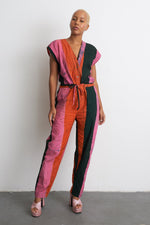Vibrant Accra Jumpsuit in orange, pink, and green hues with short sleeves and waist tie, paired with beige open-toed heels.