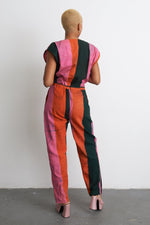 Back view of colorful Accra Jumpsuit with bold stripes, cinched waist, and short sleeves against a white wall.