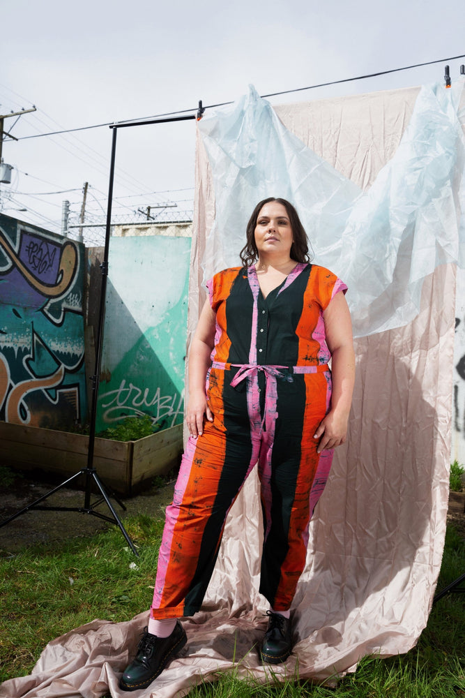 Vibrant sleeveless top and pants in orange, black, and purple, with pink belt, outdoor with graffiti art backdrop.