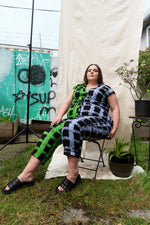 Accra Jumpsuit in Off Grid pattern, seated outdoors on chair, with pixelated black, white, and green design.