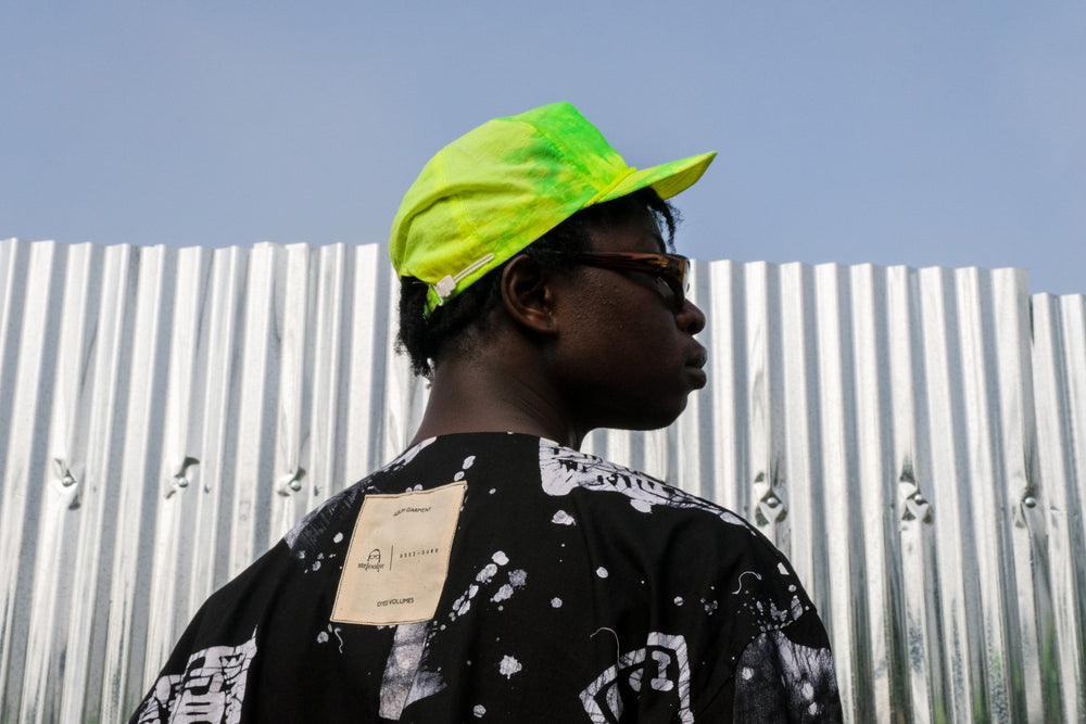 Back view of person in Album Garment shirt with white splatter pattern and green cap, against a rusted metal wall.