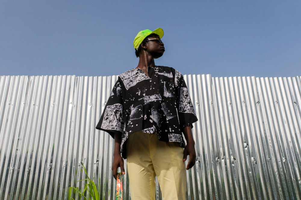 Vibrant Album Garment top, yellow trousers, green cap, in front of a corrugated metal wall under blue sky.