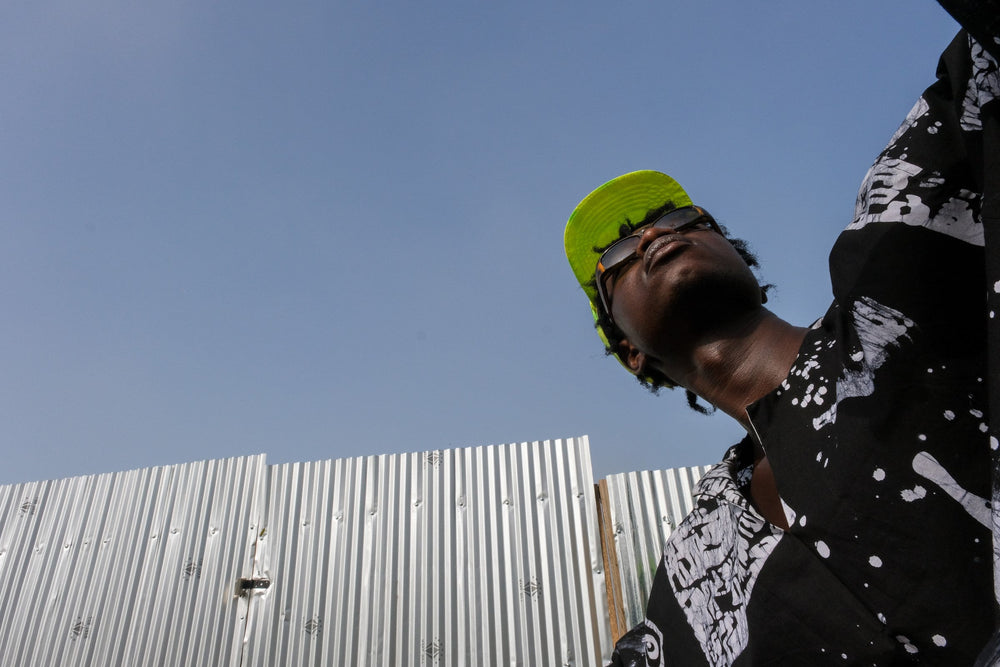 Person in Album Garment shirt looks upwards, green cap on head, against a clear sky and metal wall.