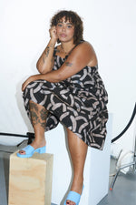 Amplo Dress in chic black & beige, seated pose on wooden block, highlighting tattoos and blue open-toed shoes.