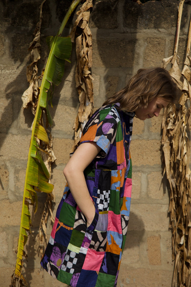 Colorful Bata Dress in Motley, sleeveless knee-length, with multicolored patchwork design, against a stone wall.