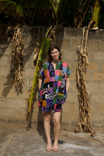 Colorful Bata Dress in Motley, short-sleeve with patchwork design, light flip-flops, against concrete block wall.