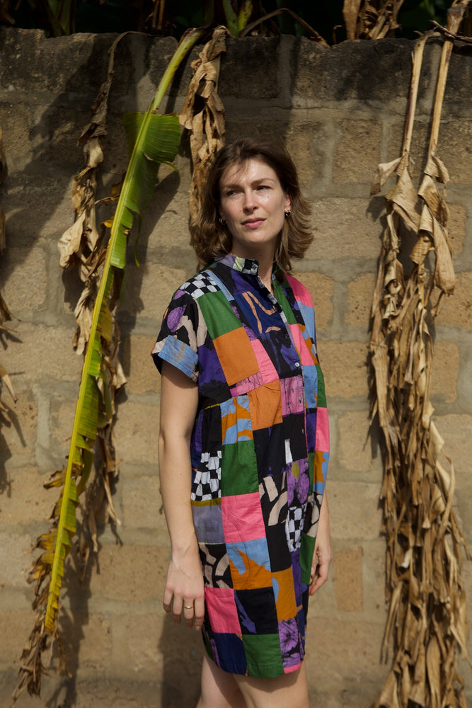 Colorful Bata Dress in Motley, short sleeves, vibrant patchwork, standing outdoors with corn stalks backdrop.