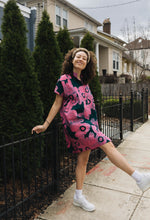 Person in pink Bata Dress strides on sidewalk, white sneakers, by black fence, green bushes, and houses on cloudy day.