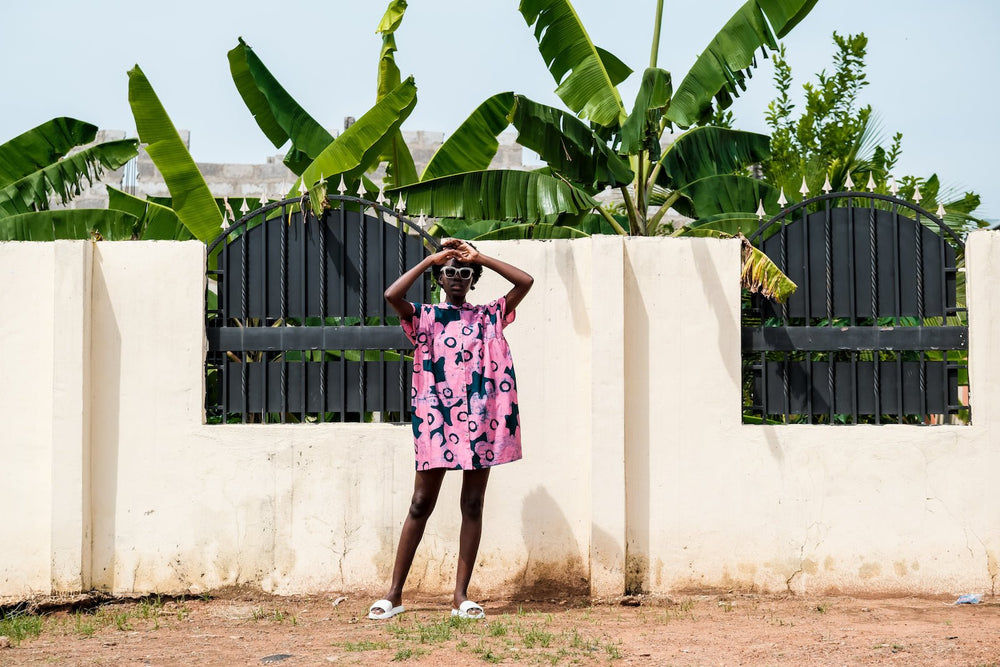 Bata Dress in pink with blue print, white sandals, hands on head, against white wall with green foliage and banana trees.