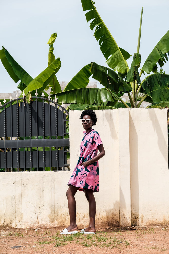 Person in pink and blue Bata Dress, white shoes, standing before a white wall with metal bars and tropical greenery.