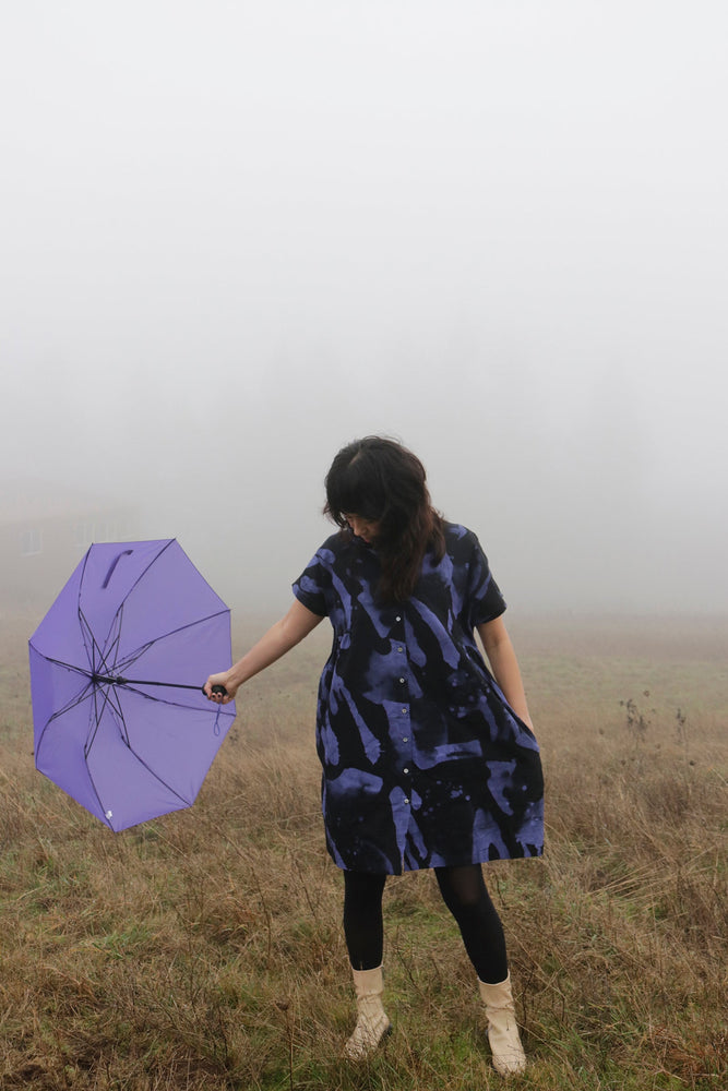 Serene Bata Dress in foggy field, purple umbrella in hand, showcasing unique print and style.
