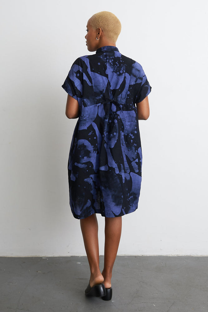 Back view of Bata Dress with tie waist, ethereal abstract blueish-purple print, in front of a white wall.