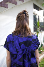 Blueish-purple and black Bata Dress, model has medium-length hair, outdoor setting with white structure.