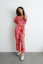 Person in vibrant Tee Hee Easy Jumpsuit with abstract batik print, tied waist, paired with white shoes, against a white wall.