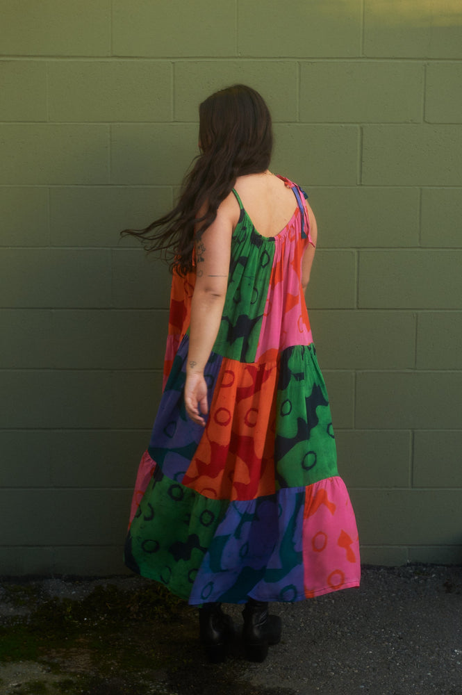 Back veiew of the colorful Esca Dress in Cool Cool Cool, ankle-length with thin straps and dark boots, against a green wall.