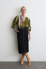 Stylish Flos Blouse with green, purple, yellow stripes, black skirt, and tan boots, against a white wall.