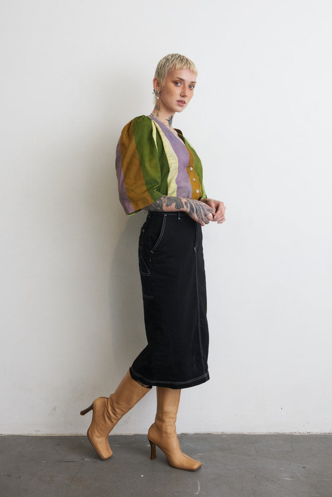 Vibrant Flos Blouse in green and purple, paired with a black skirt and tan boots, against a white wall.