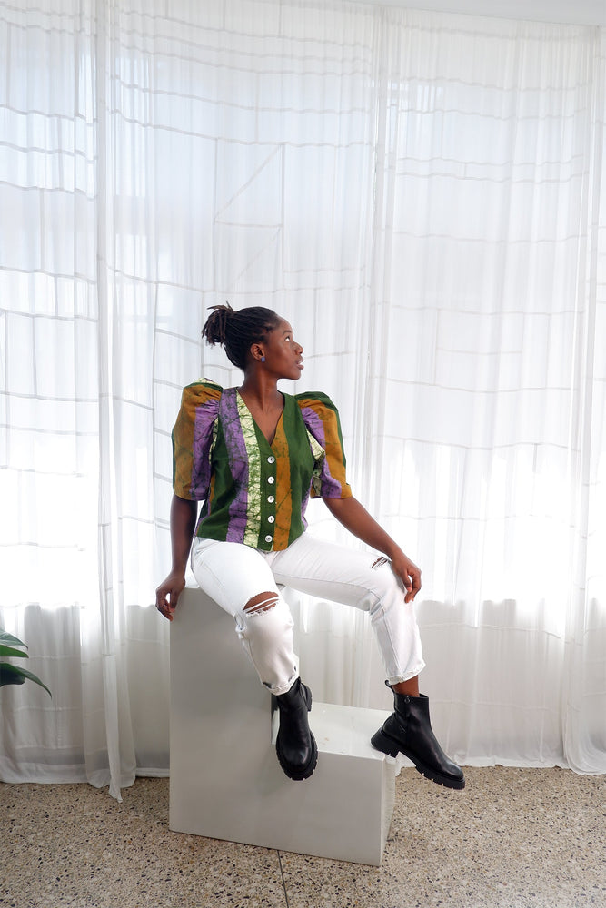 Green-dominant Flos Blouse, white ripped jeans, black boots, seated by window with plants.