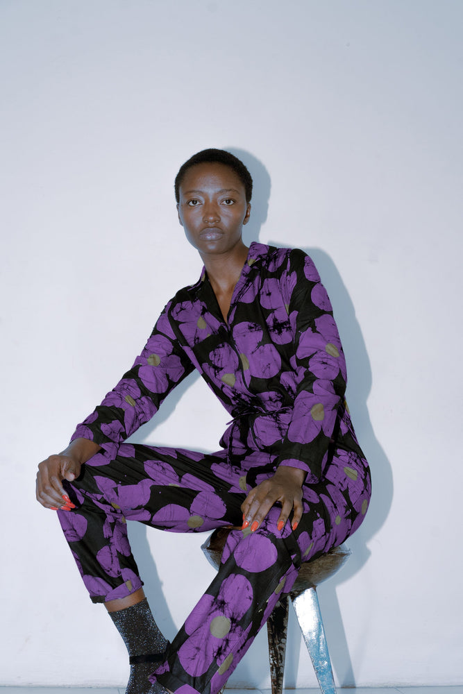 Seated model in Love Perfect floral jumpsuit, vibrant purple pattern, with red nail accent, on metallic stool.