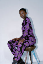 Woman in Love Perfect floral jumpsuit seated on rustic stool, large purple flowers on dark cotton fabric.