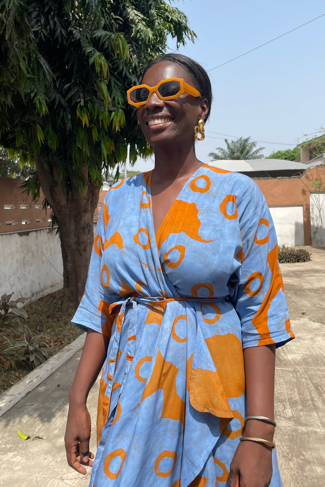 A woman standing outside in a blue and orange dress and sunglasses, looking stylish in the Letsa Wrap Dress in All Ideas.