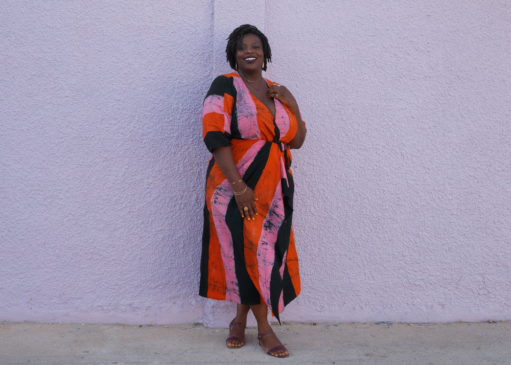 Colorful Letsa wrap dress in orange, dark green, and pink print, paired with open-toed sandals, against a textured wall.
