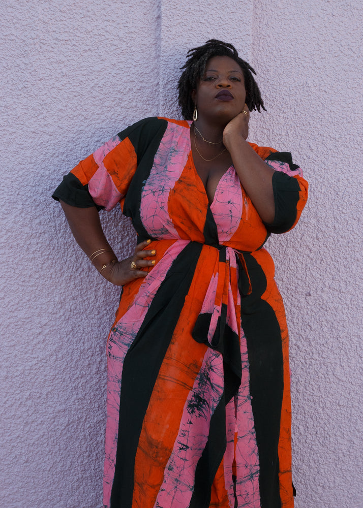 Colorful Letsa wrap dress in Carmine print, tied waist, model poses with hand on neck against a textured backdrop.