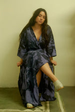 Seated individual in Letsa dress with open front, blue-black print, white slippers, and minimalist chair.