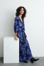 Flowing Letsa dress with abstract blueish purple print, tied waist, and black boots, beside a white pedestal.