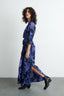 Blueish purple and black Letsa dress with brush stroke pattern and black boots, mid-step against a white wall and grey floor.
