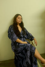 Person seated on chair in blue and black patterned Letsa dress in Rorschach, against a plain light-colored wall.