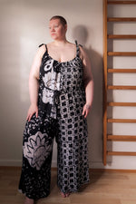Ligo Jumpsuit in 2 Party System print, featuring adjustable tie straps and a loose fit, worn by model standing next to a ladder.