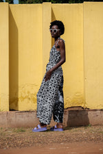 Side view of the sleeveless Ligo Jumpsuit in black and white pattern, model wears sunglasses and purple slippers against yellow wall.