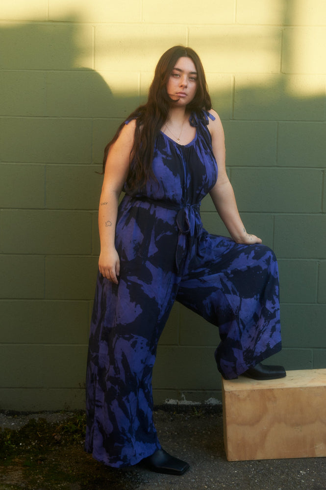 Ligo Jumpsuit in Rorschach, highlighting the loose fit, adjustable tie straps, and optional sash against a green backdrop.