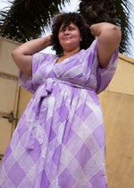 Person in purple Eostre print dress with waist tie, hands on head, standing by palm tree, embodying relaxed elegance.