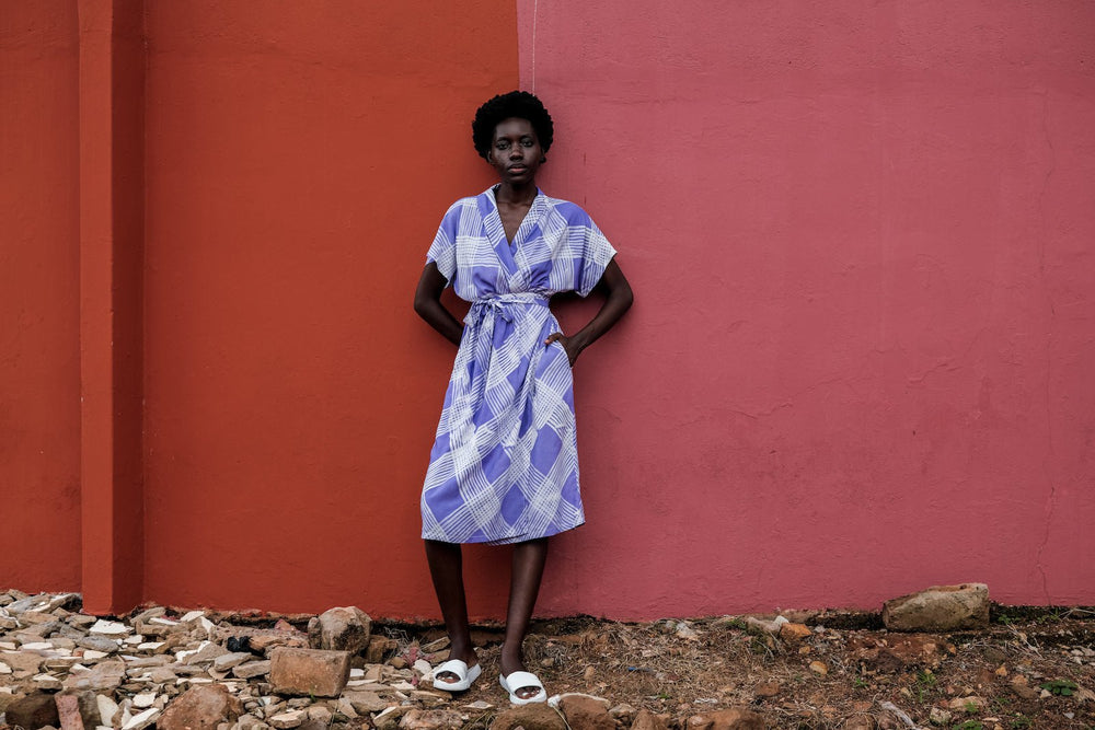 Person in Sampa Dress against red wall, showcasing the dress’s white and purple print and casual white sandals.