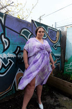 Tea-length Sampa Dress in front of graffiti art, purple and white stripes, blending urban backdrop with soft femininity.